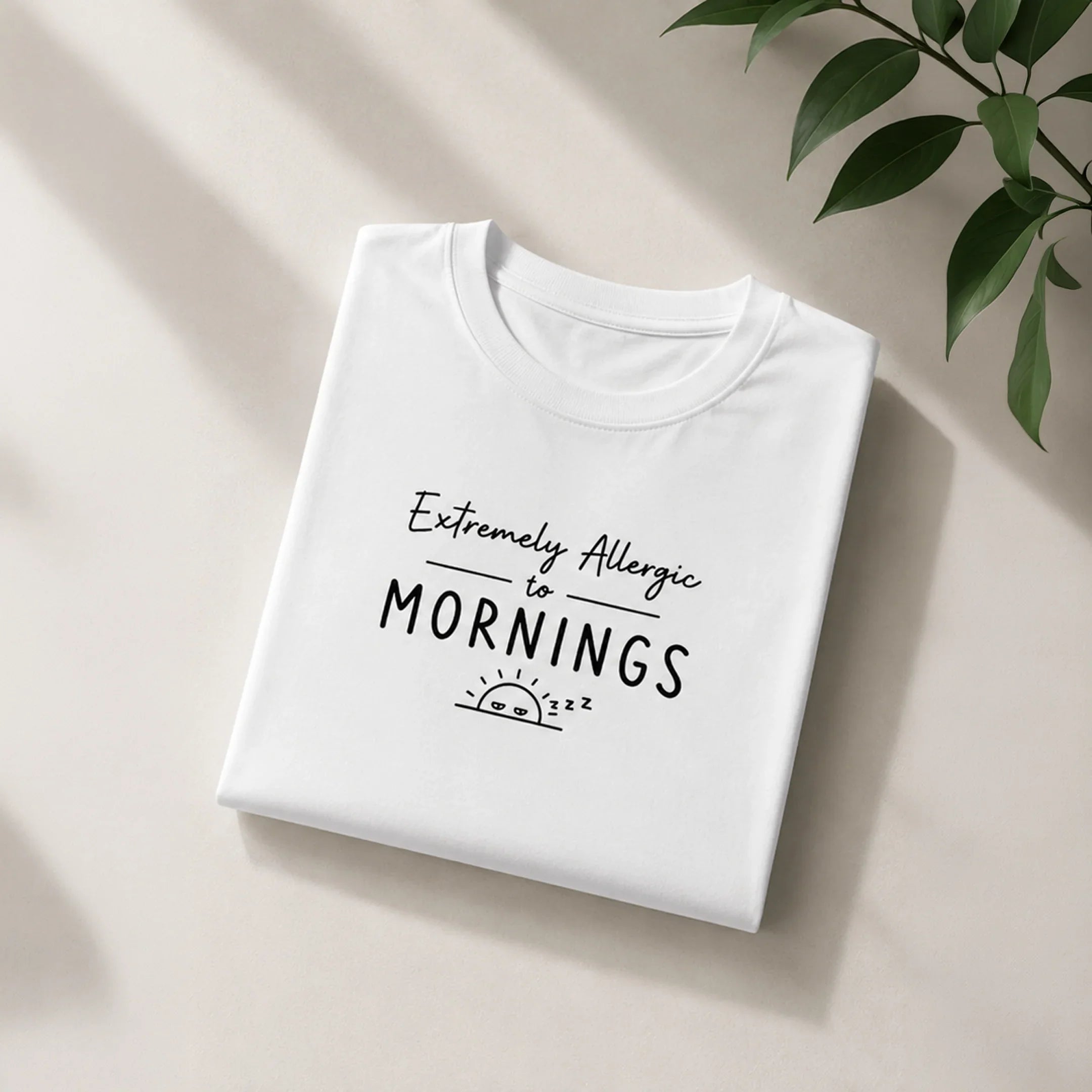 White graphic tee with 'Extremely Allergic to Mornings' text and sleepy sun design, styled with green leaves.