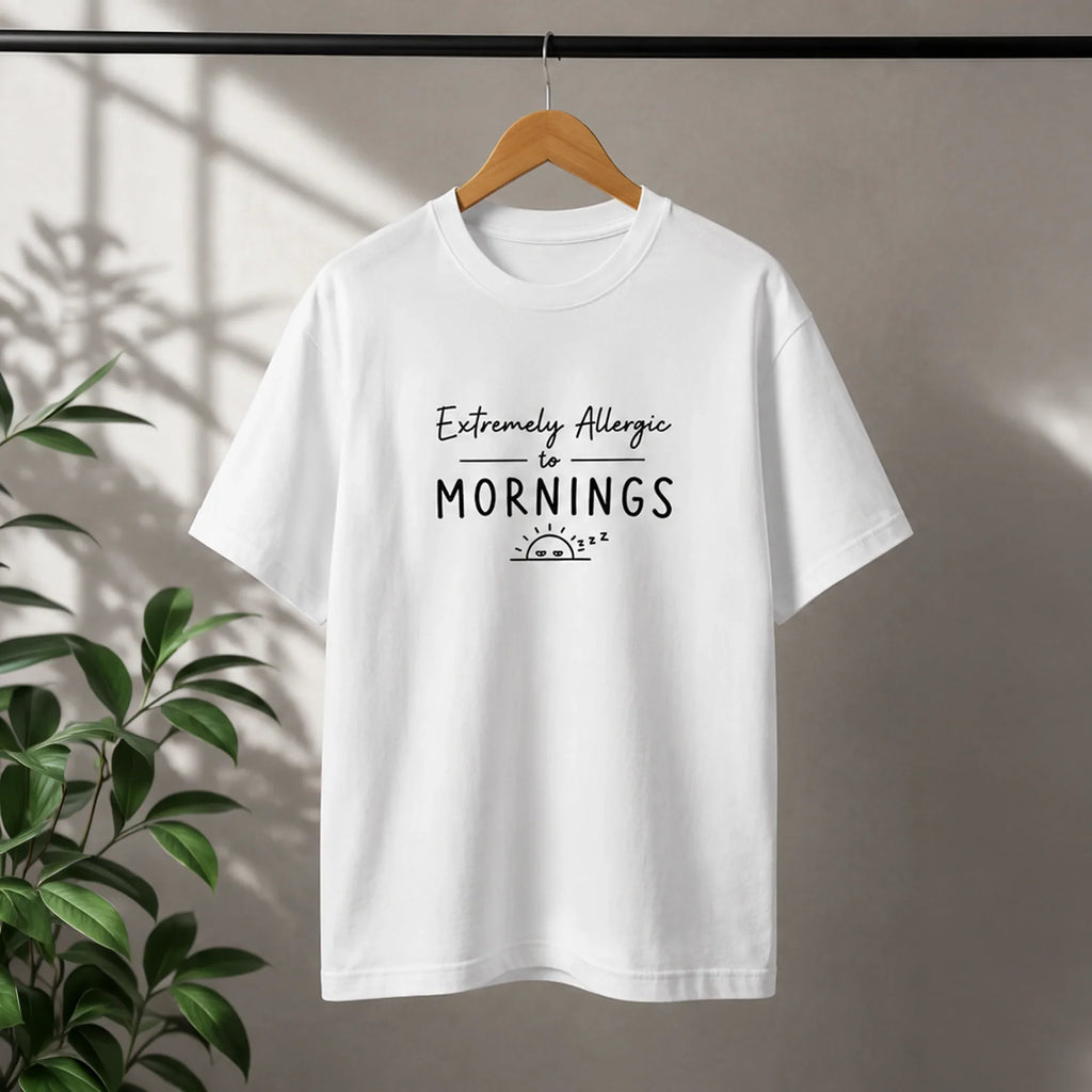 White graphic tee with 'Extremely Allergic to Mornings' text and sleepy sun design, hanging on wood hanger