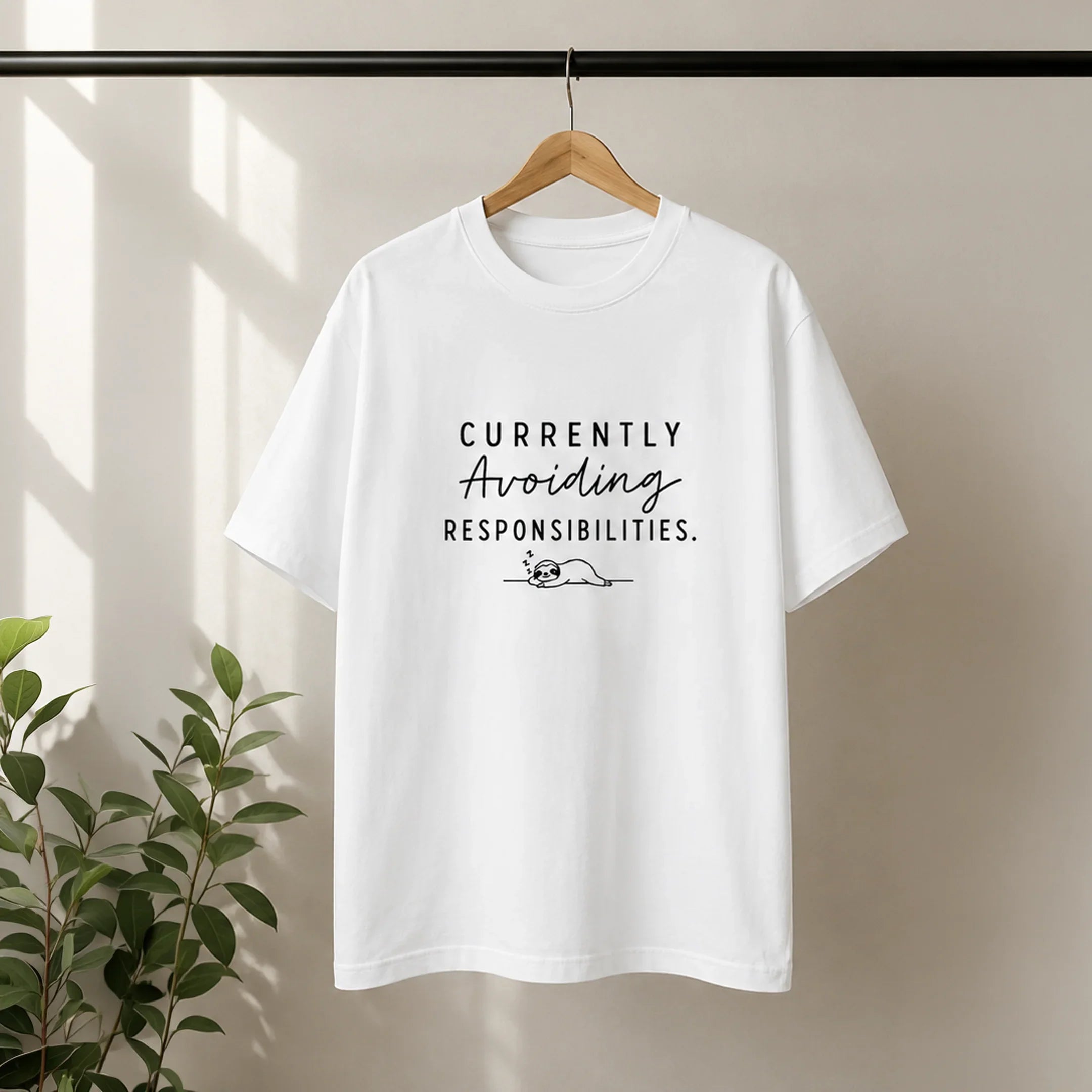 White graphic tee with 'Currently Avoiding Responsibilities' text and sloth design, hanging on hanger